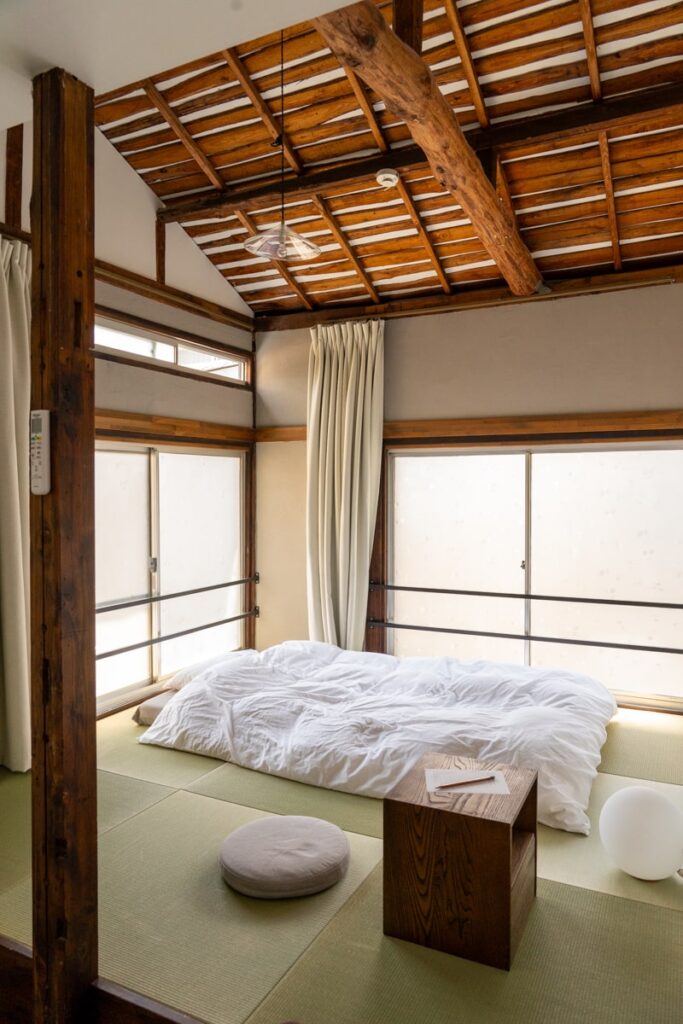 A minimalist traditional Japanese room with a futon bed on tatami flooring, illuminated by natural light.