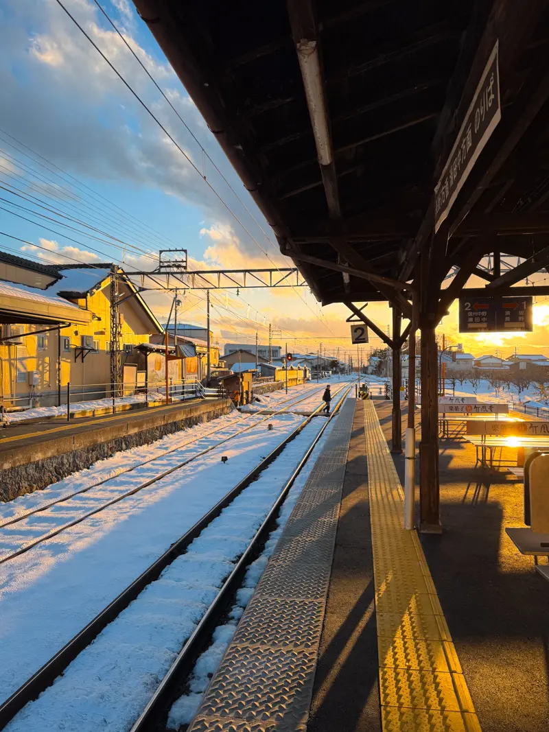 A quiet train platform (Obuse Station) bathed in golden sunlight, with a snowy mountain landscape in the distance in Nagano, Japan.